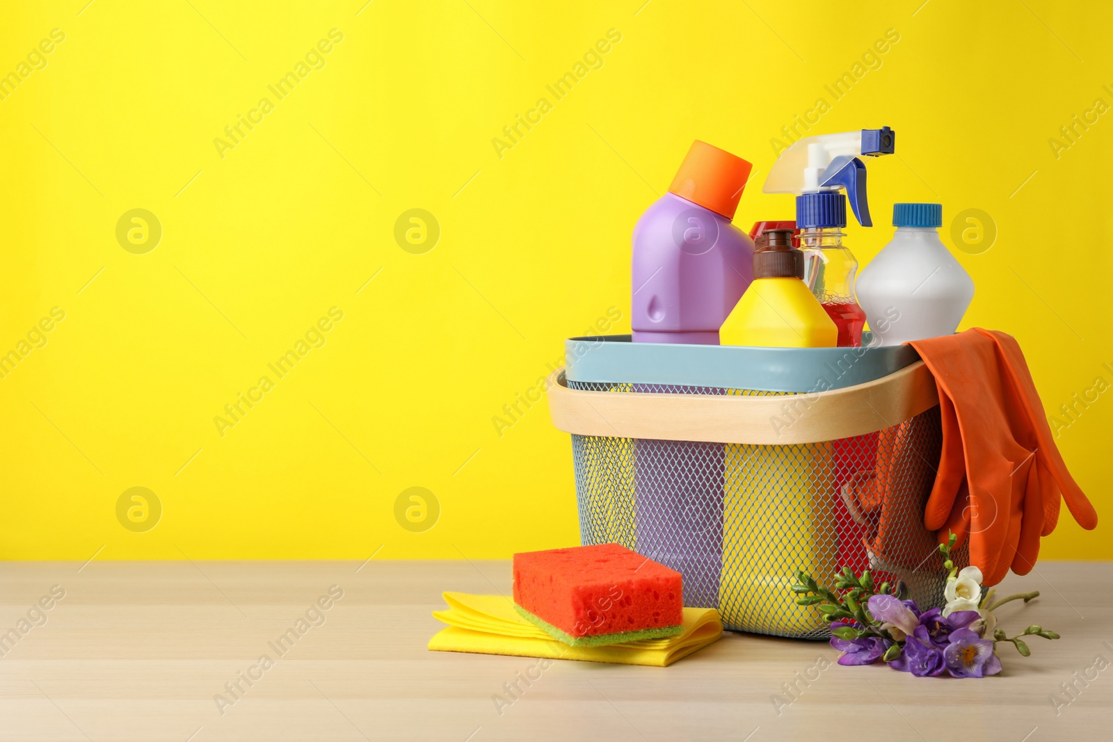 Photo of Plastic basket with spring flowers and cleaning supplies on wooden table. Space for text