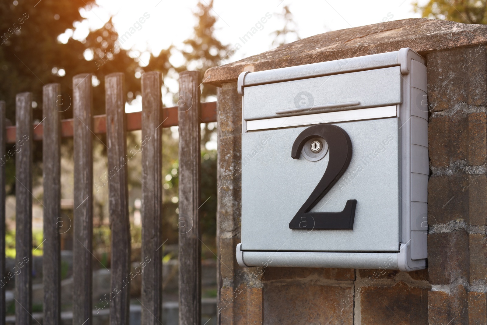 Photo of Metal mailbox with number 2 on stone fence near wooden gate outdoors, space for text