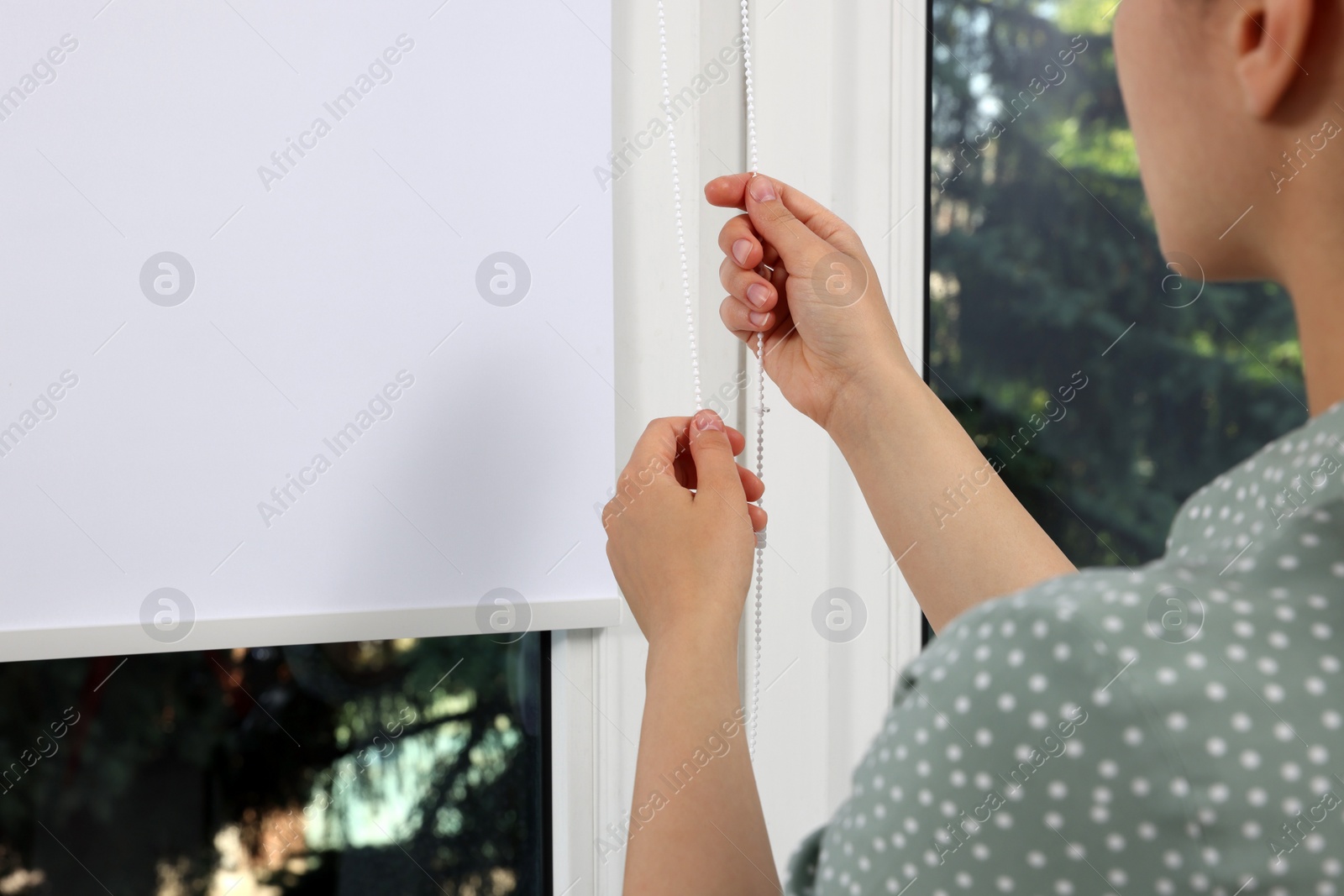 Photo of Woman opening white roller blind on window indoors, closeup