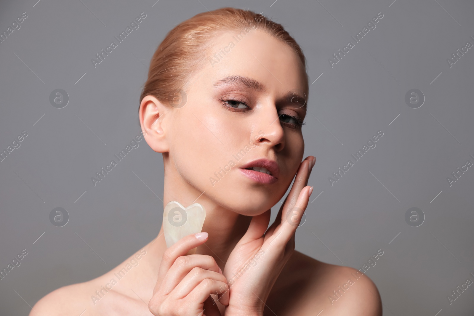 Photo of Beautiful young woman doing facial massage with gua sha tool on grey background, closeup