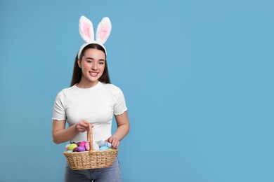 Photo of Happy woman in bunny ears headband holding wicker basket of painted Easter eggs on turquoise background. Space for text