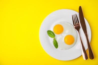 Photo of Tasty fried eggs with basil in plate on yellow background, top view. Space for text