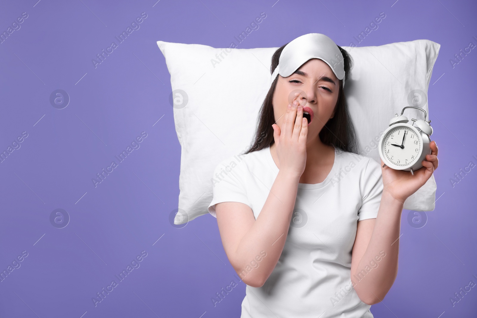 Photo of Tired young woman with sleep mask, alarm clock and pillow on purple background, space for text. Insomnia problem