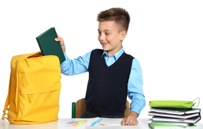 Photo of Little boy in uniform putting book into backpack at desk against white background. School stationery