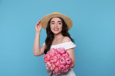 Beautiful young woman in straw hat with bouquet of pink peonies against light blue background