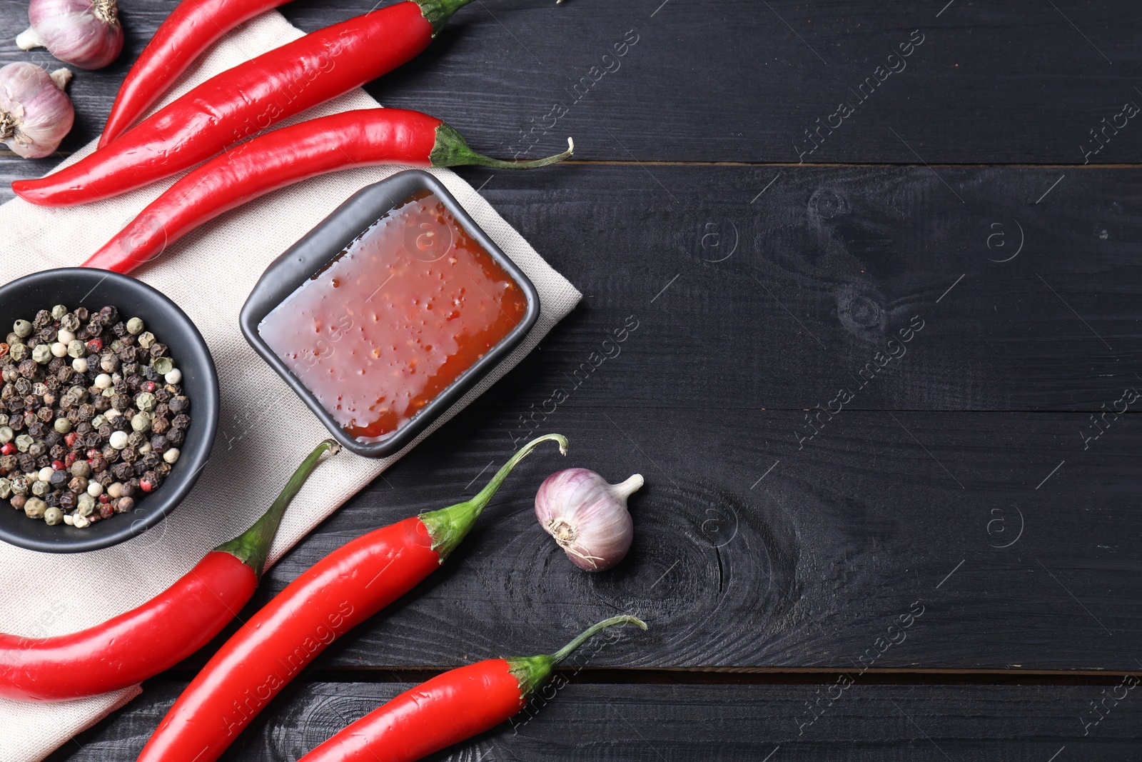 Photo of Spicy chili sauce, garlic, peppers and peppercorns on black wooden table, flat lay. Space for text