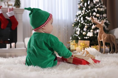 Baby in cute elf costume near Christmas gift on floor at home, back view