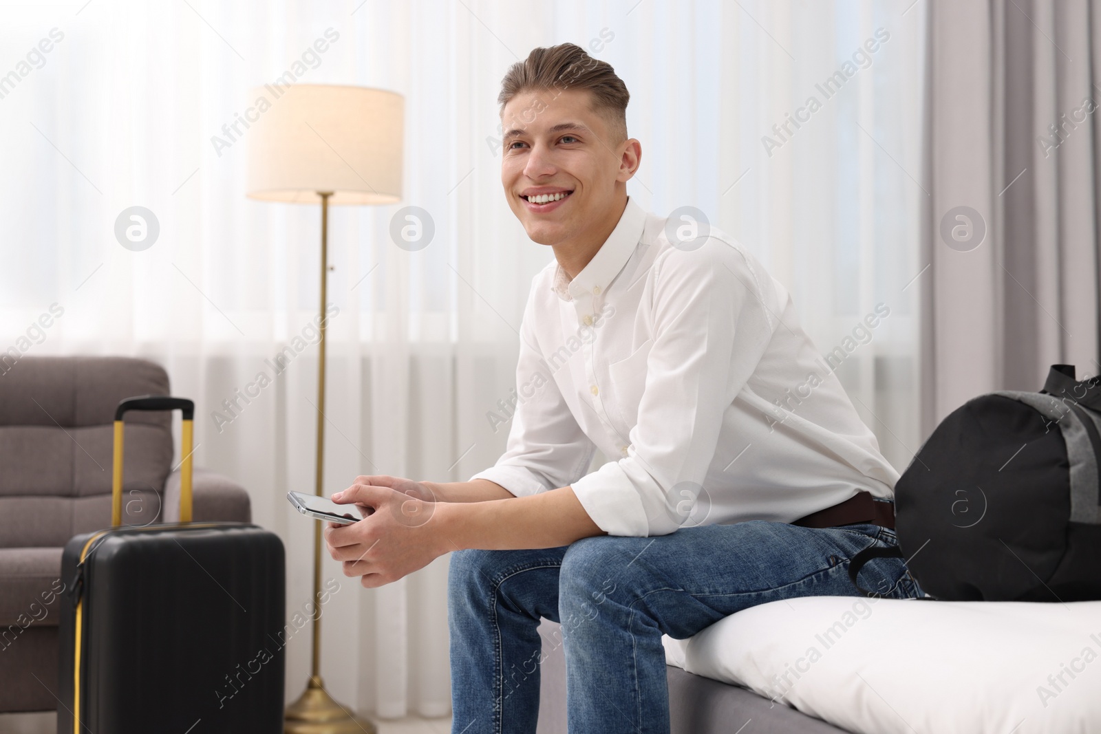 Photo of Smiling guest with smartphone relaxing on bed in stylish hotel room