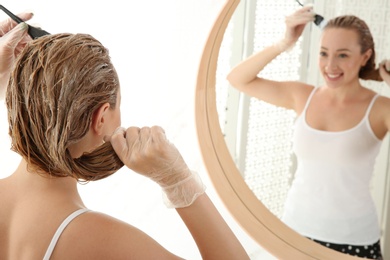 Photo of Young woman dyeing her hair in front of mirror at home