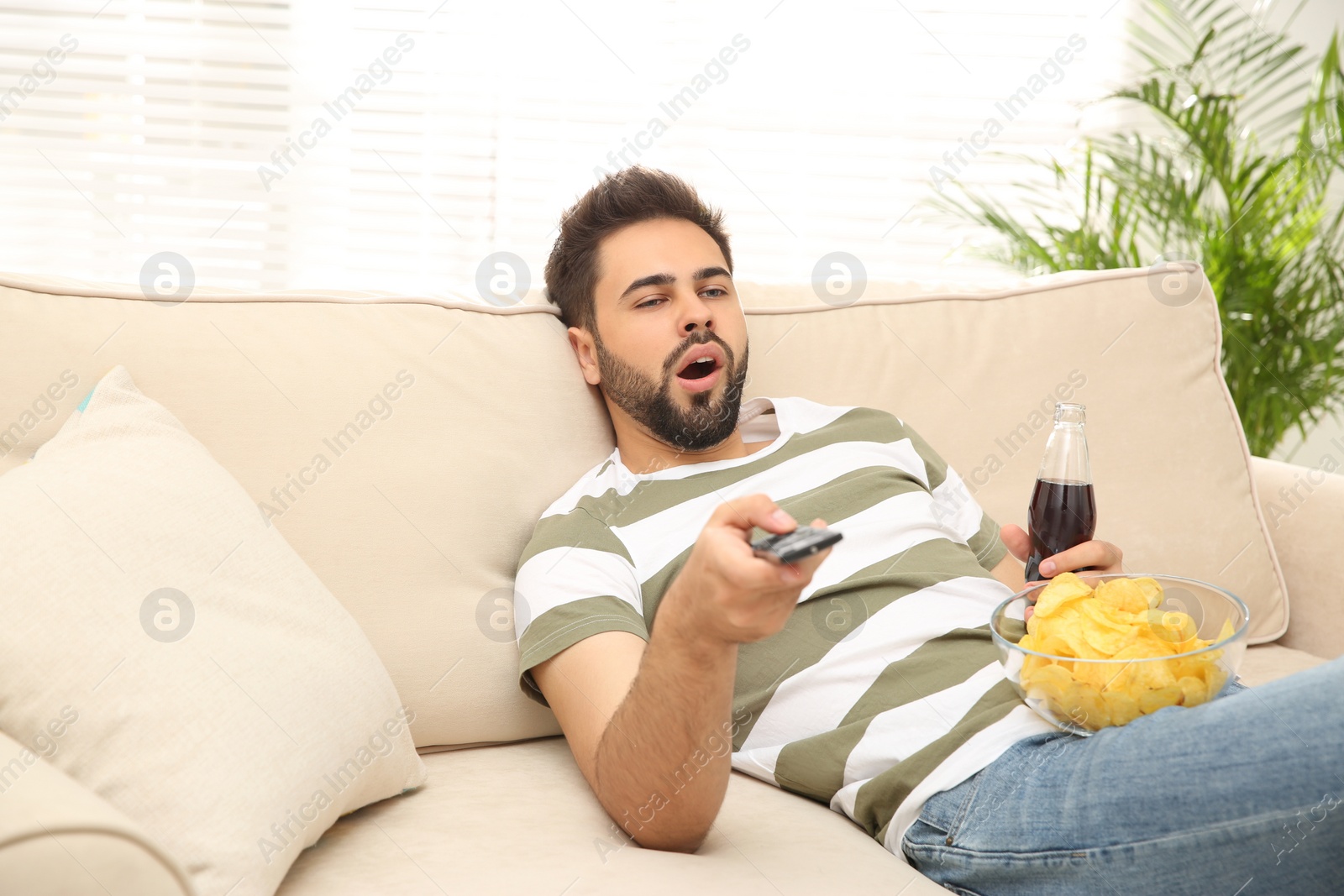 Photo of Lazy young man with chips and drink watching TV on sofa at home