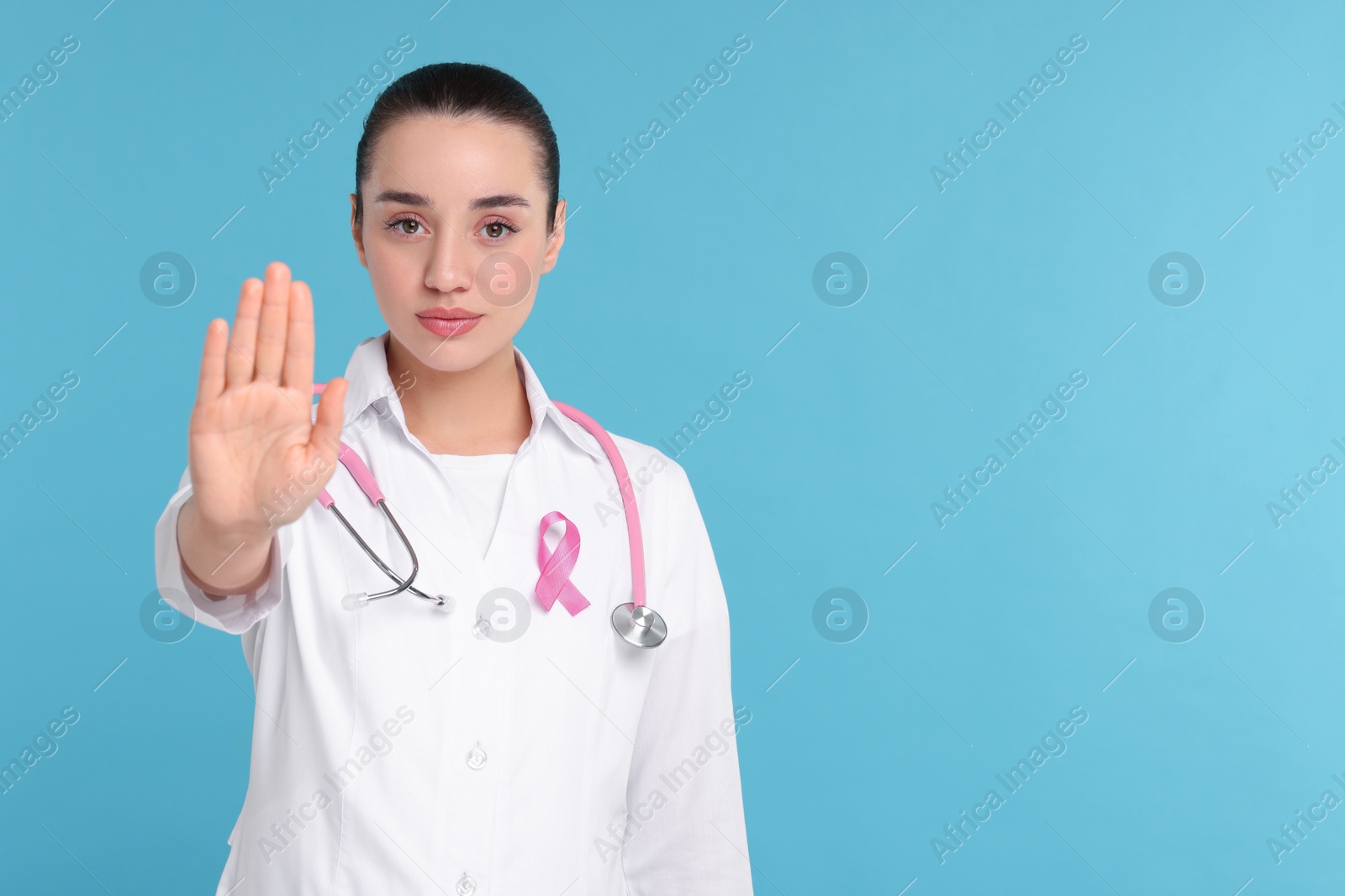 Photo of Mammologist with pink ribbon showing stop gesture on light blue background, space for text. Breast cancer awareness