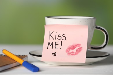 Photo of Sticky note with phrase Kiss Me, lipstick mark, cup of drink and notebook on white table against blurred background, closeup