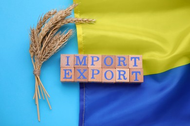 Photo of Words Import and Export made of wooden cubes, ears of wheat and Ukrainian flag on light blue background, flat lay