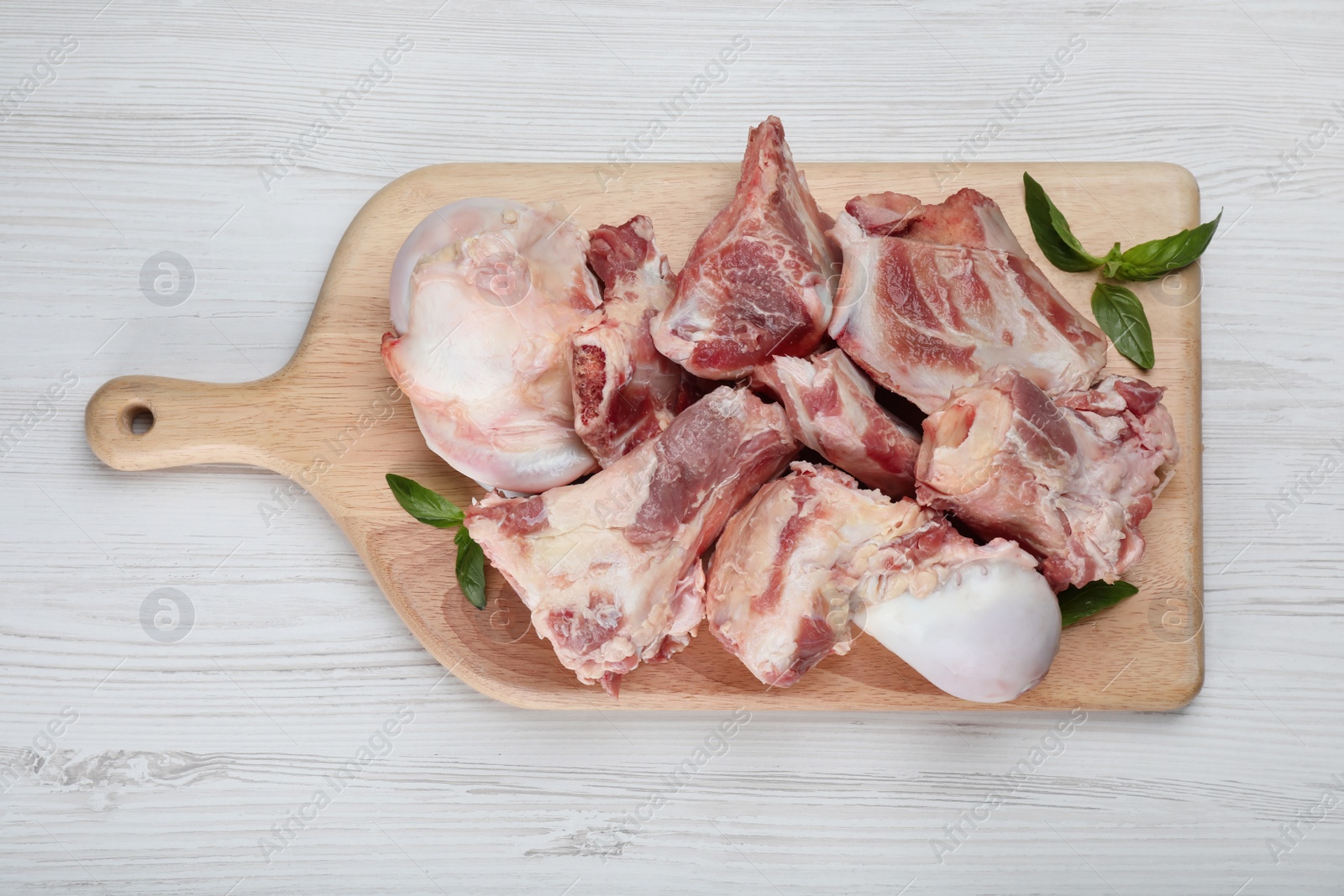 Photo of Cutting board with raw chopped meaty bones and basil on white wooden table, top view