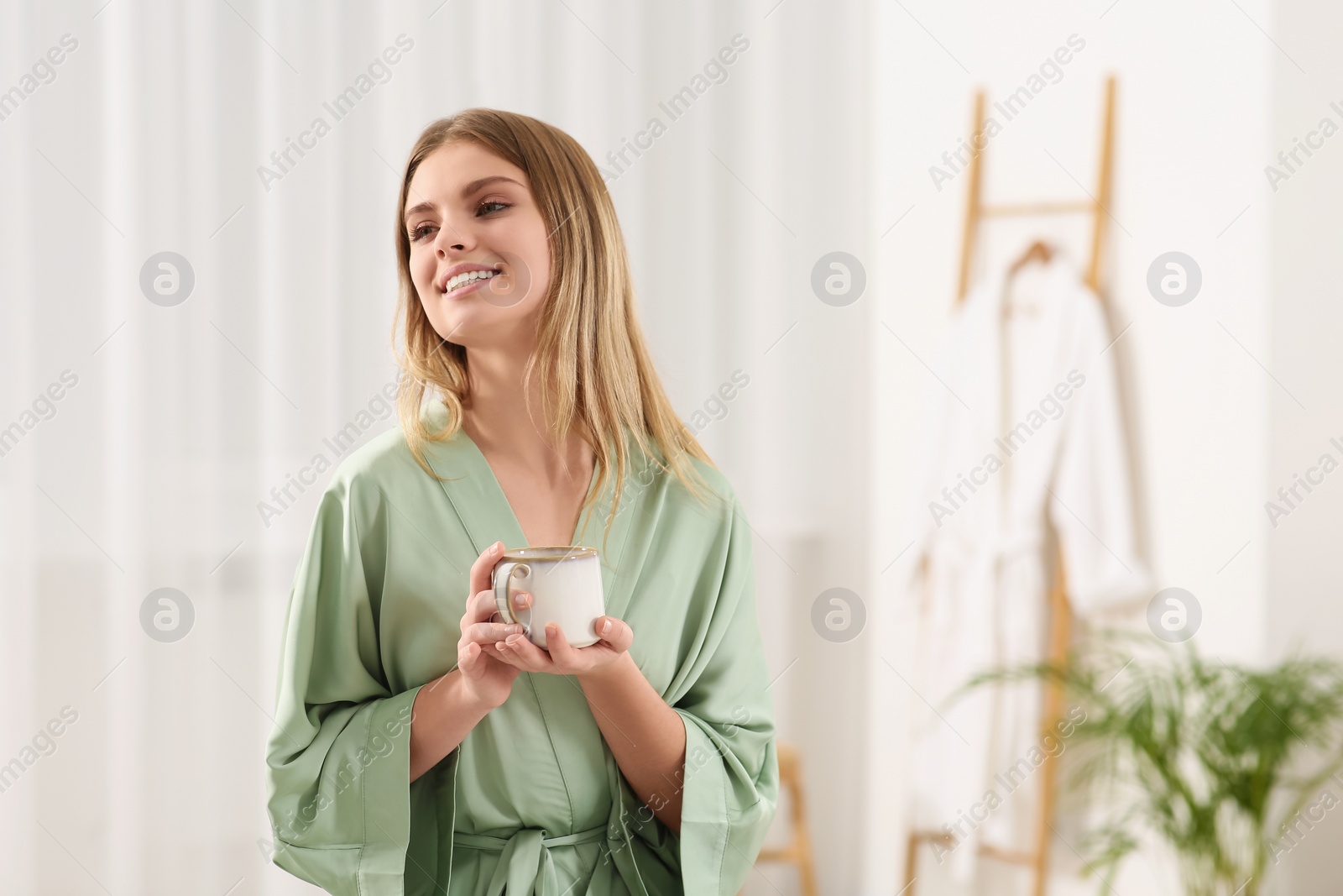 Photo of Beautiful happy woman with cup of hot drink wearing robe in bathroom. Space for text