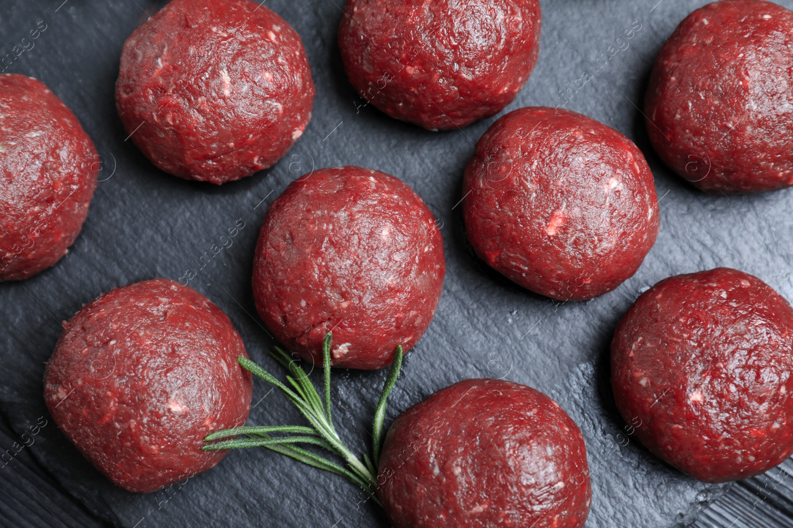 Photo of Many fresh raw meatballs with rosemary on black board, flat lay