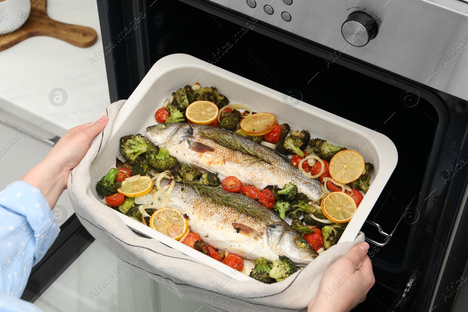 Photo of Woman taking baking dish with delicious fish and vegetables from oven in kitchen, closeup