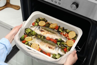 Photo of Woman taking baking dish with delicious fish and vegetables from oven in kitchen, closeup