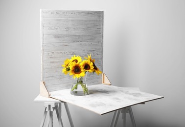 Photo of Glass vase with beautiful sunflowers and double-sided backdrop on table in photo studio