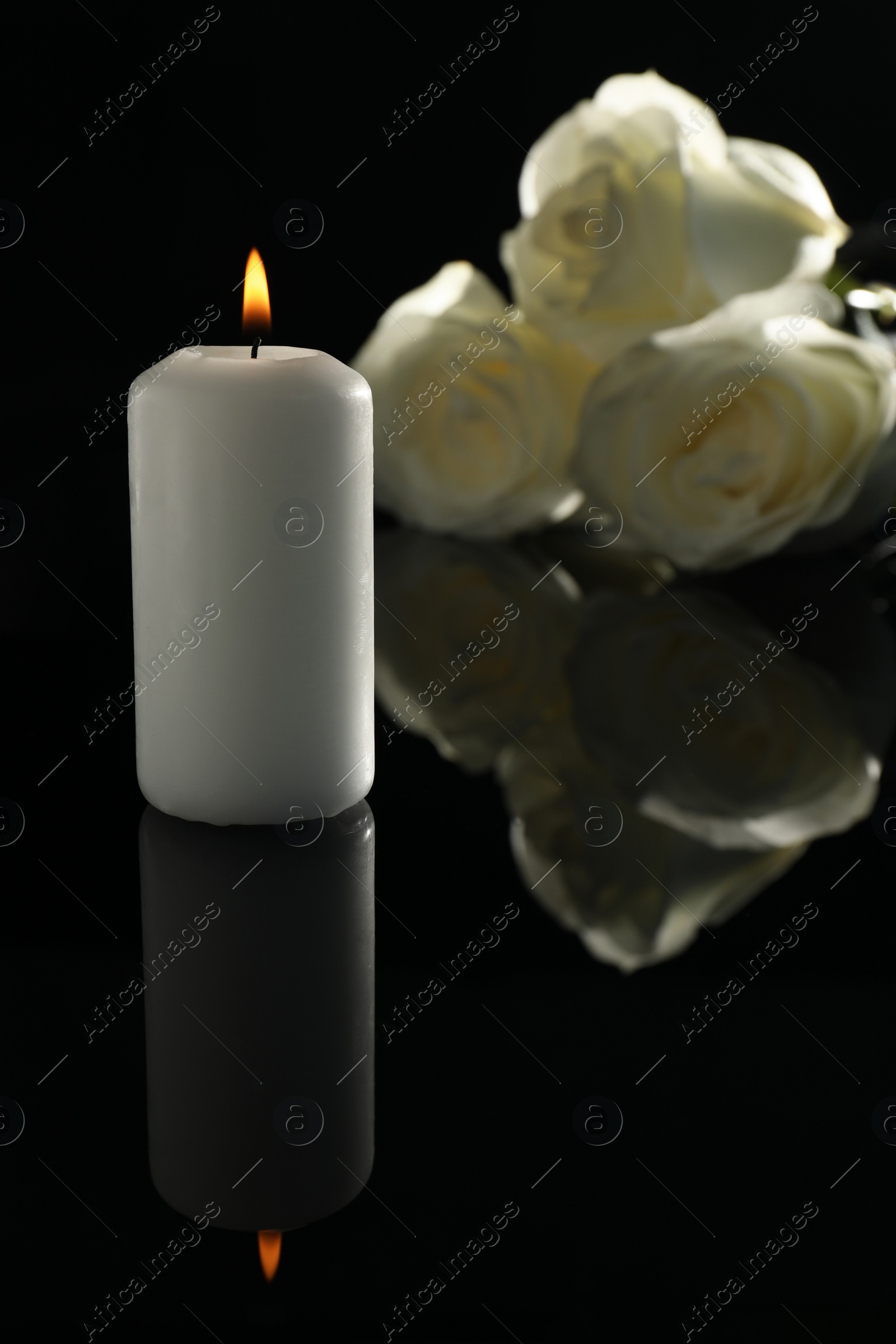 Photo of White roses and burning candle on black mirror surface in darkness. Funeral symbols