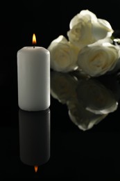 White roses and burning candle on black mirror surface in darkness. Funeral symbols