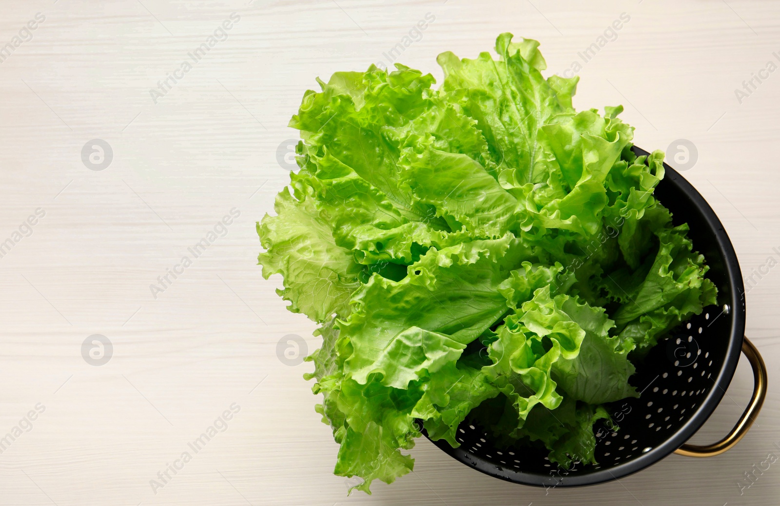 Photo of Fresh lettuce in black colander on white wooden table, top view. Space for text