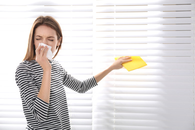 Photo of Young woman suffering from dust allergy while cleaning window blinds at home