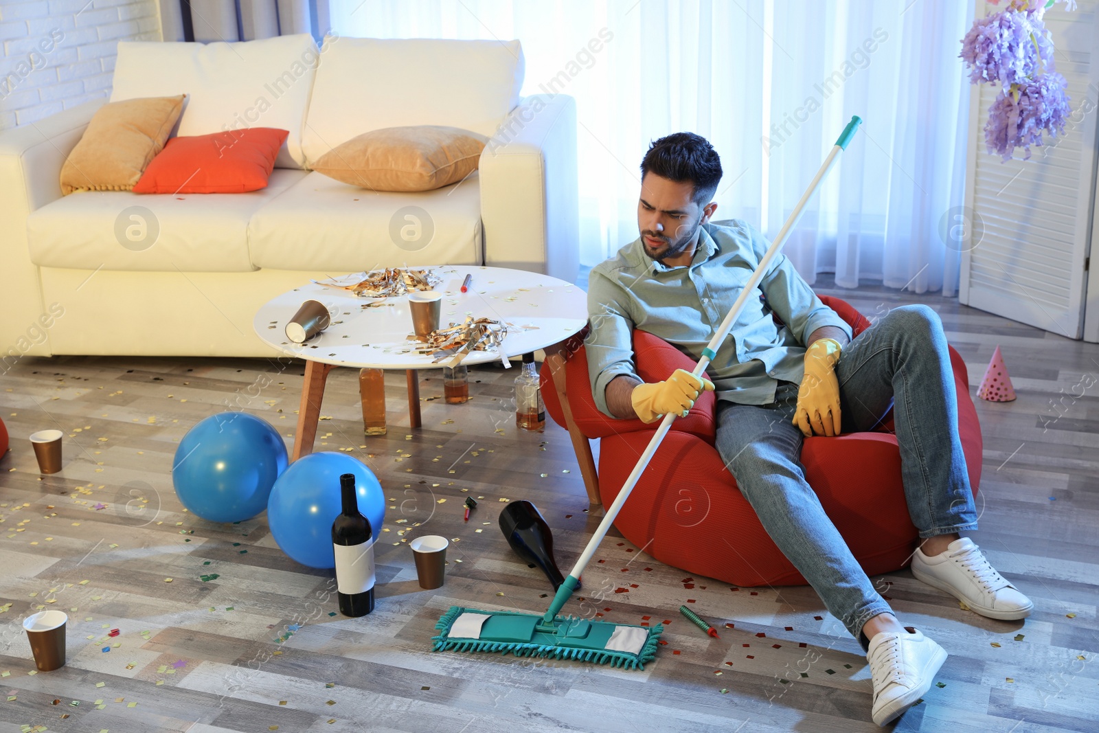 Photo of Young man with mop suffering from hangover in messy room after party