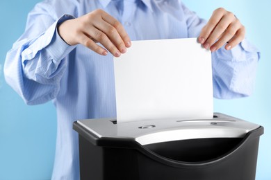Photo of Woman destroying sheet of paper with shredder on light blue background, closeup
