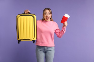 Emotional young woman with passport, ticket and suitcase on purple background