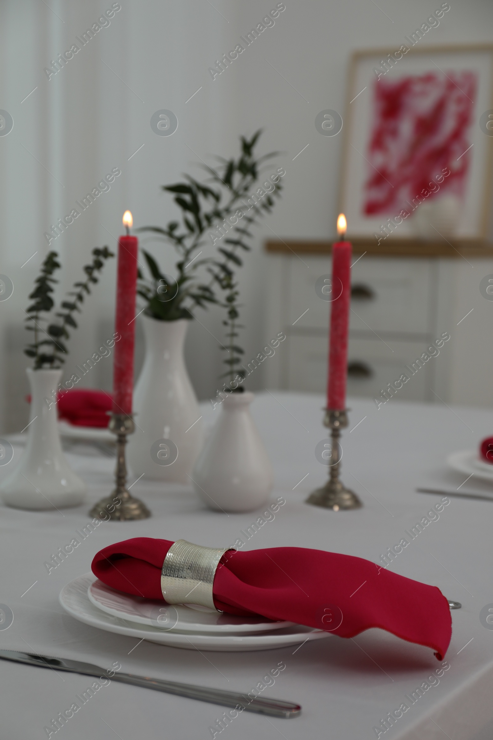 Photo of Table setting. Plates, pink napkin, cutlery, burning candles and vases with green branches in dining room