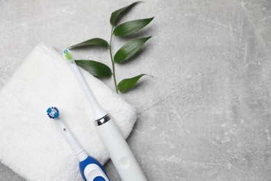 Electric toothbrushes, towel and leaves on grey textured table, flat lay. Space for text