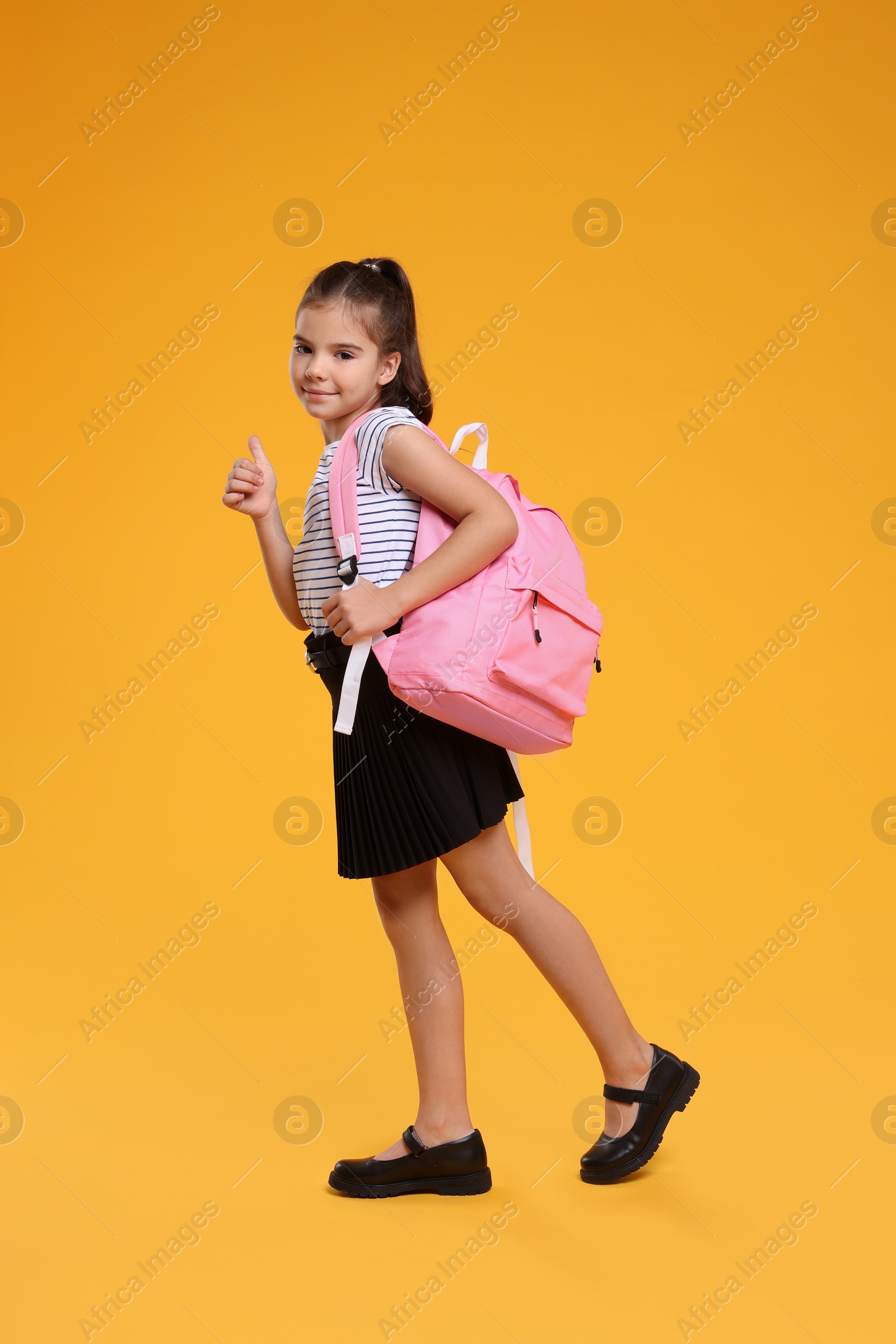 Photo of Back to school. Cute girl with backpack walking and showing thumbs up on orange background