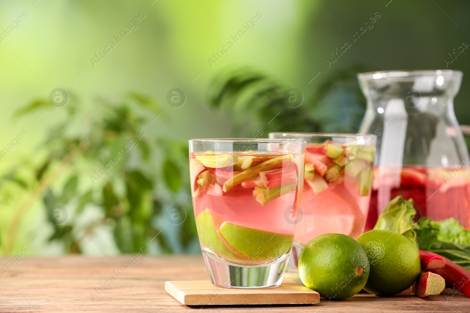 Photo of Glasses and jug of tasty rhubarb cocktail with lime fruits on wooden table outdoors, closeup. Space for text