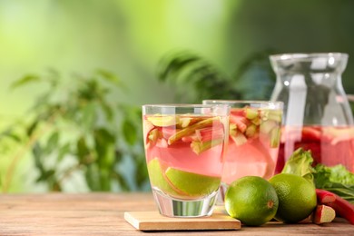 Photo of Glasses and jug of tasty rhubarb cocktail with lime fruits on wooden table outdoors, closeup. Space for text