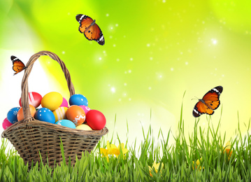 Image of Easter eggs in wicker basket on green grass and butterflies against blurred background