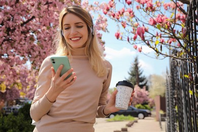 Photo of Happy woman with smartphone and coffee listening to audiobook outdoors on spring day