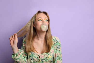 Photo of Fashionable young woman with bright makeup blowing bubblegum on lilac background, space for text