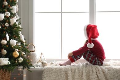 Photo of Cute little girl in Santa hat on window sill near Christmas tree at home