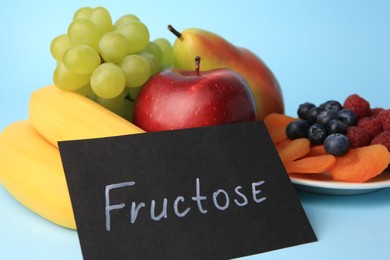 Photo of Card with word Fructose, delicious ripe fruits, berries and dried apricots on light blue background, closeup