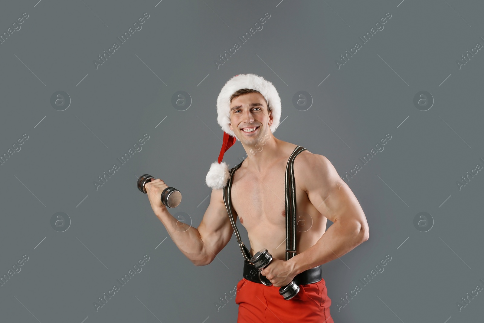 Photo of Young muscular man in Santa hat with dumbbells on gray background