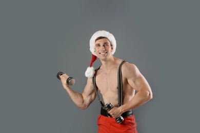 Photo of Young muscular man in Santa hat with dumbbells on gray background