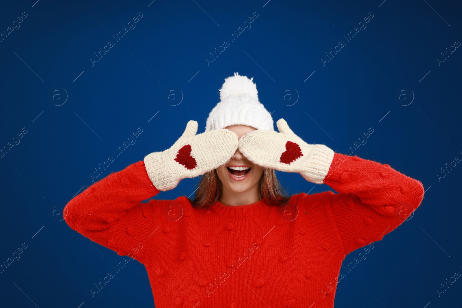 Photo of Funny young woman in warm sweater, mittens and hat on blue background. Winter season