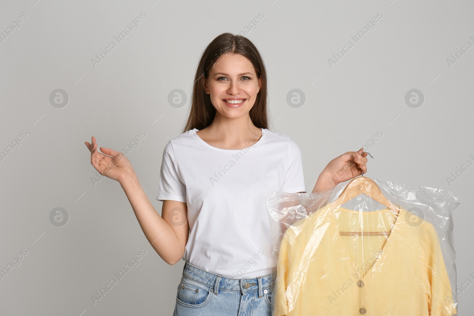 Photo of Young woman holding hanger with blouse in plastic bag on light grey background. Dry-cleaning service