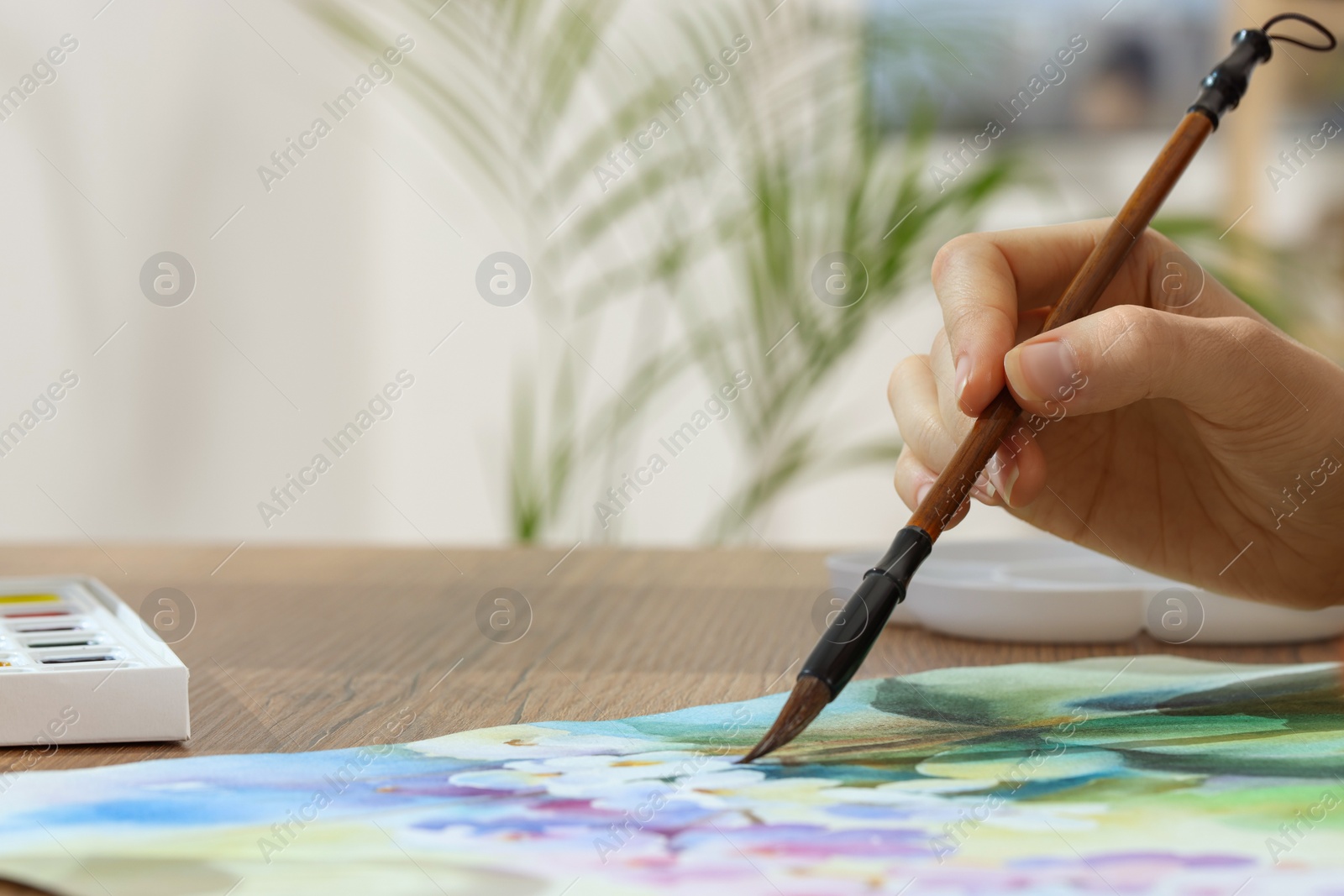 Photo of Woman painting flowers with watercolor at wooden table, closeup. Space for text