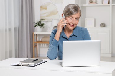 Beautiful senior woman talking on phone while using laptop at white table indoors. Space for text
