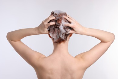 Young woman washing her hair with shampoo on light grey background, back view