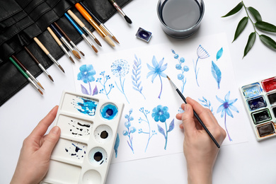 Woman painting flowers with watercolor at white table, top view