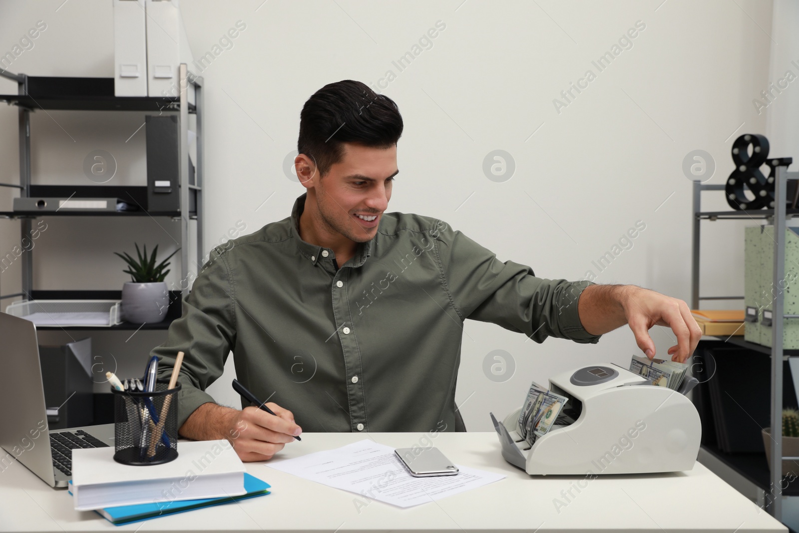 Photo of Man putting money into banknote counter at white table indoors