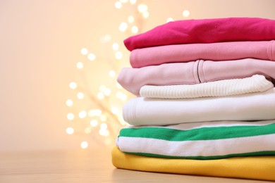 Photo of Stack of folded clothes on wooden table against beige background with blurred lights, space for text